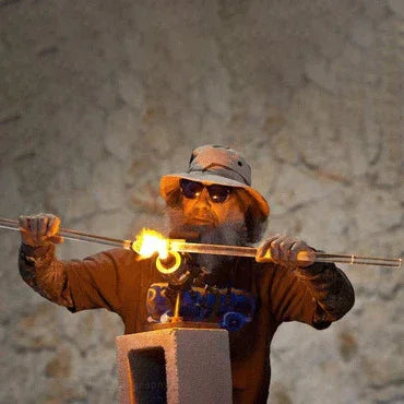 Glass artist in sunglasses shaping heated glass tube with torch in studio setting
