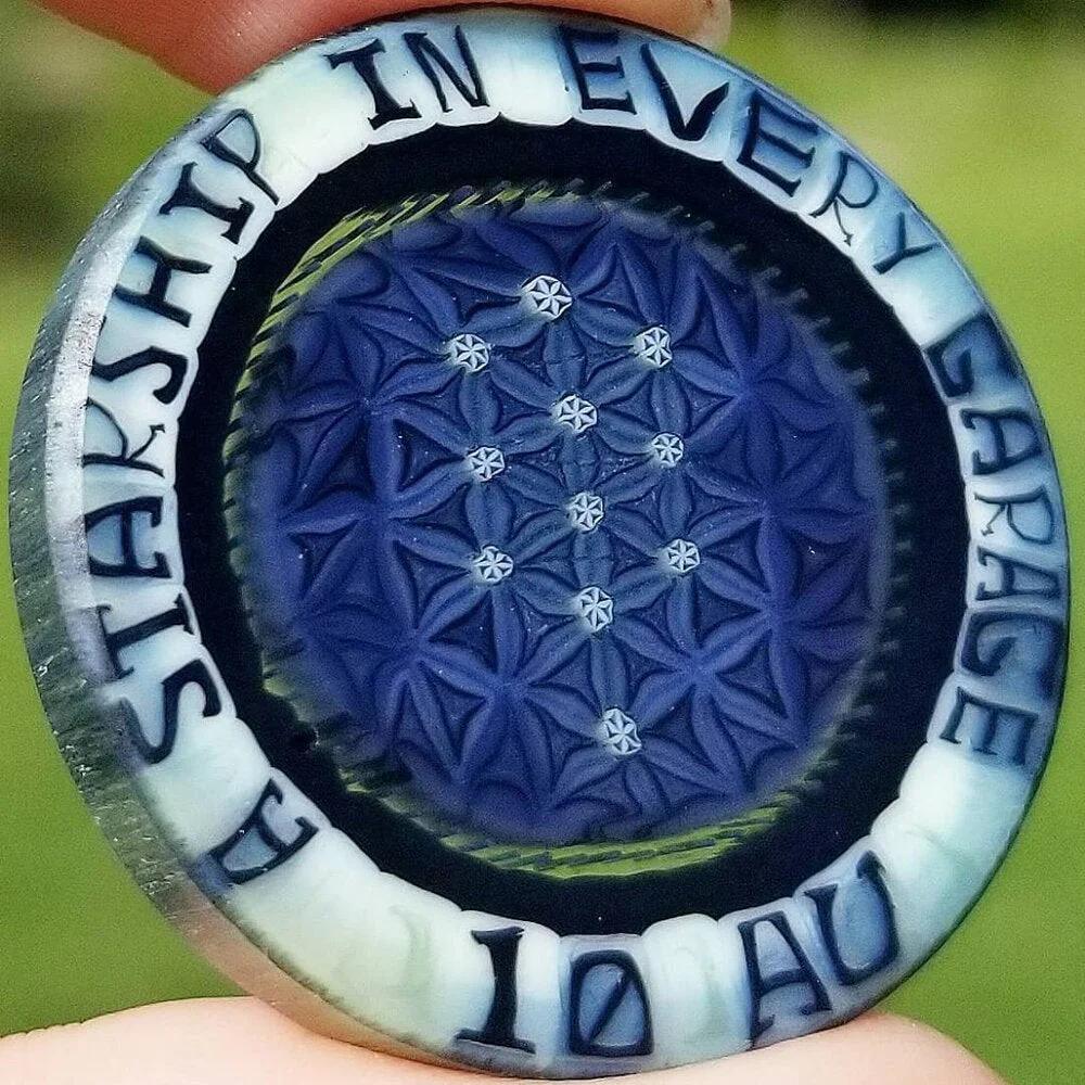 Close-up of a blue glass coin pendant with geometric patterns and engraved text 'A STARSHIP IN EVERY GARAGE 10 AU'.