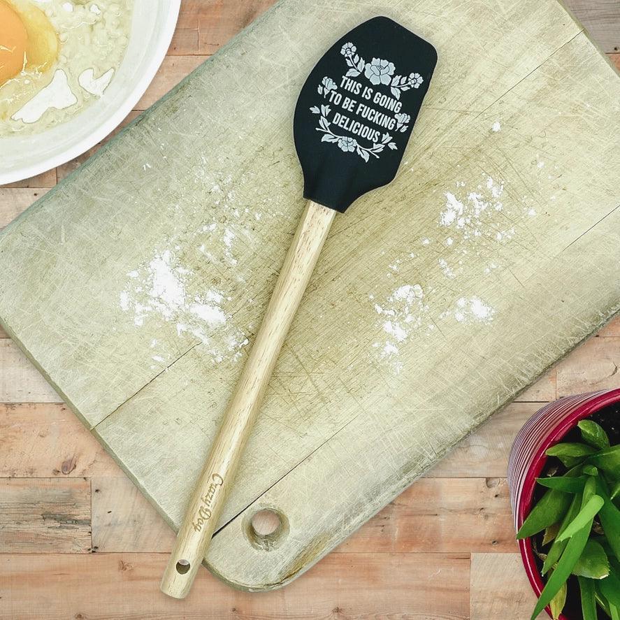 Black silicone spatula with humorous text on wooden cutting board next to mixing bowl and plant.