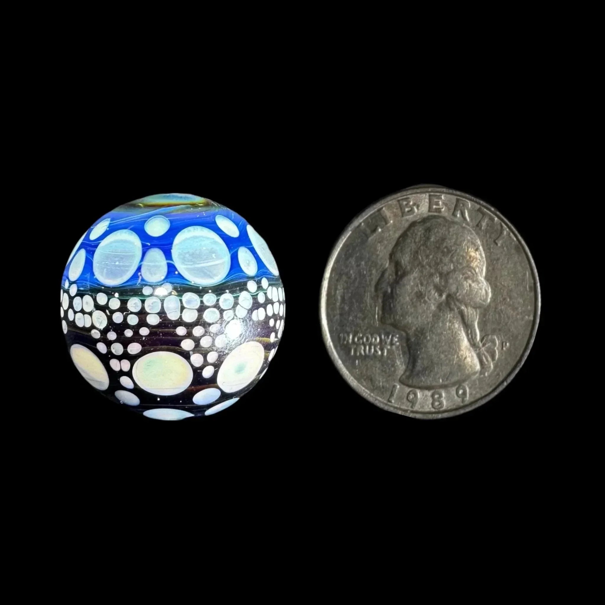 Colorful glass marble with blue and white dot design next to a US quarter for size comparison
