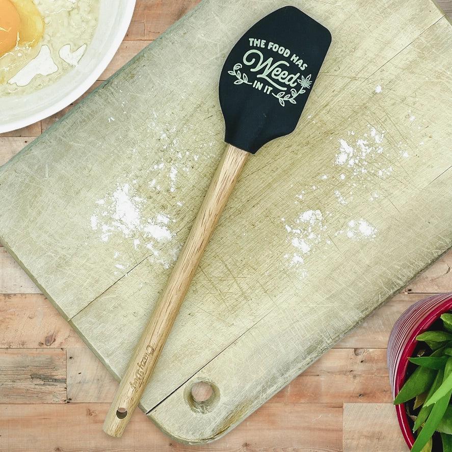 Black silicone spatula with 'The Food Has Weed In It' text on a wooden cutting board, baking scene.
