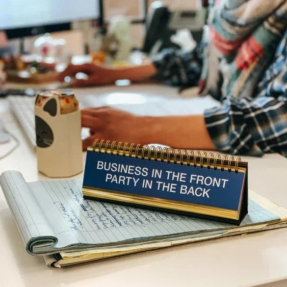 Desk scene with spiral sign reading 'Business in the front, party in the back', notebook and mug