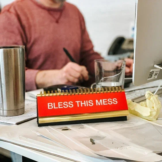 Desk with clutter, red sign reading 'Bless This Mess', tumbler, and computer monitor