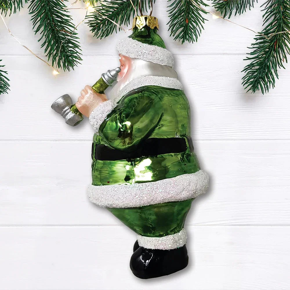 Green Santa ornament holding a glass pipe, hanging under pine branches on white wood background.