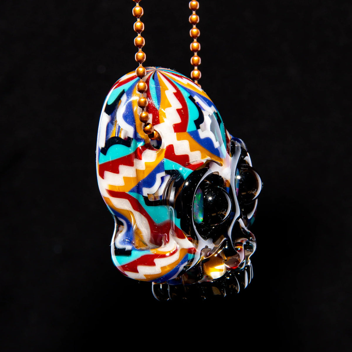 Colorful glass skull pendant with zigzag patterns on gold ball chain, against black background