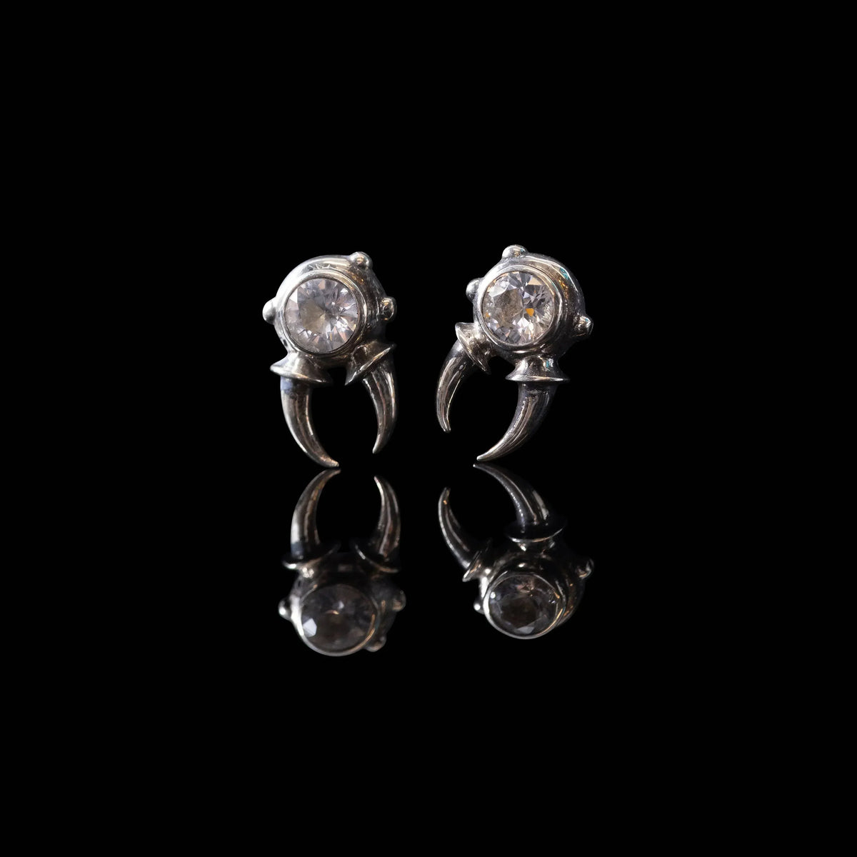 Pair of silver claw stud earrings with round clear gemstones on black reflective background