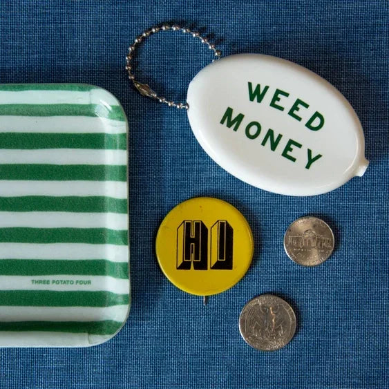 White coin pouch labeled 'Weed Money' with striped tray, yellow pin, and coins on blue background.