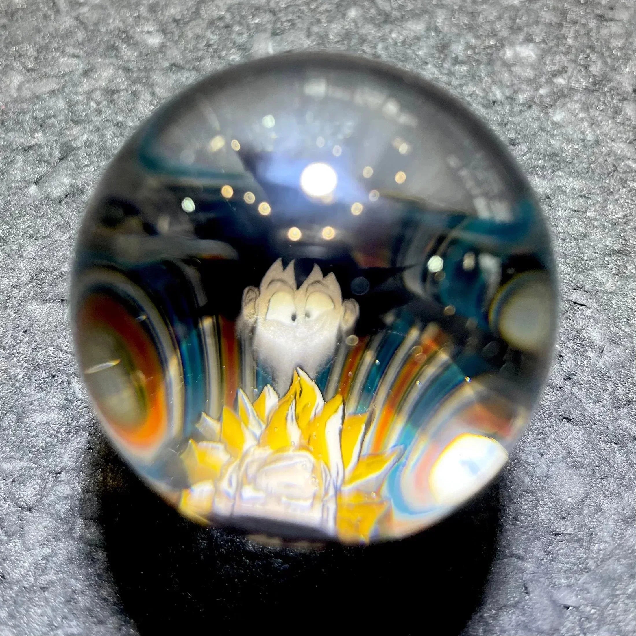 Hand-blown glass marble with anime character and colorful flame design, on textured surface