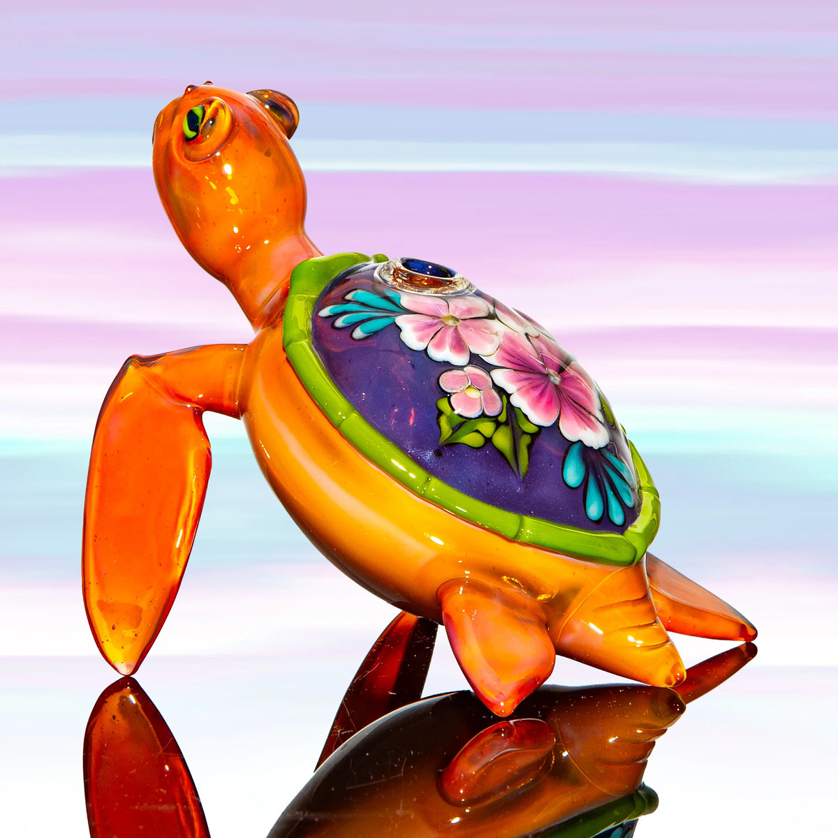 Hand-blown glass turtle pipe with colorful floral shell on reflective surface, pastel background.