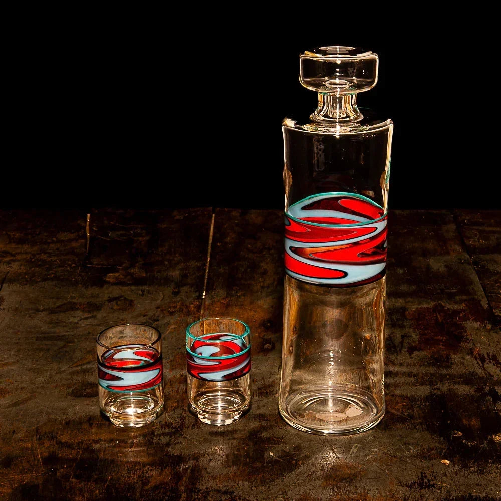 Hand-blown glass decanter and two glasses with red and blue swirl accents on wood surface