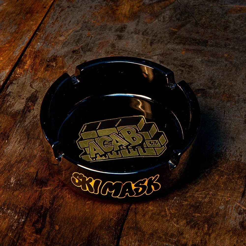 Black glass ashtray with yellow graffiti-style 'SKI MASK' and 'ACAB' text, on wood table