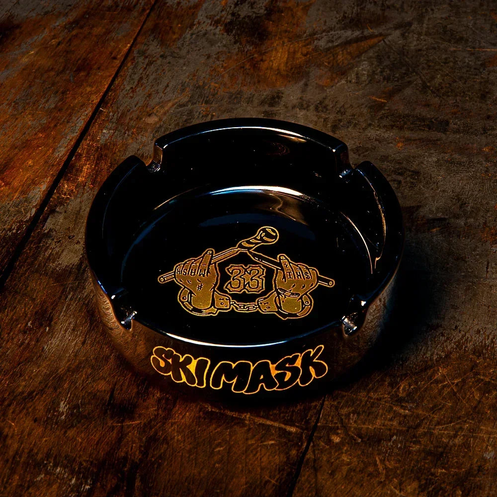Black glass ashtray with gold 'SKIMASK' lettering and handcuff graphic on rustic wood table