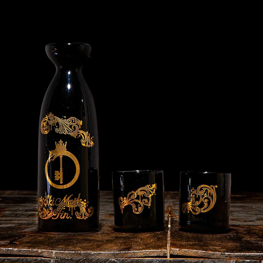 Black glass decanter and two cups with ornate gold designs on rustic surface