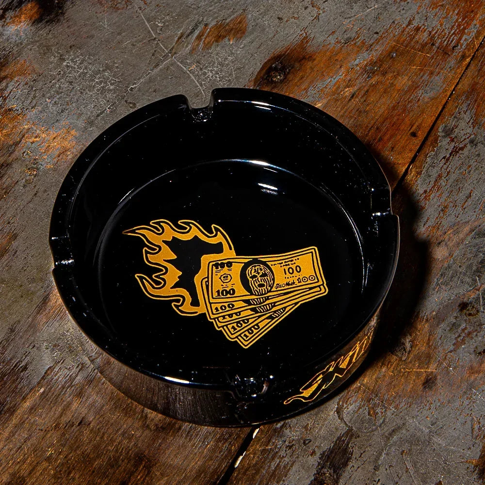 Black glass ashtray with gold flame and money graphic on rustic wooden table