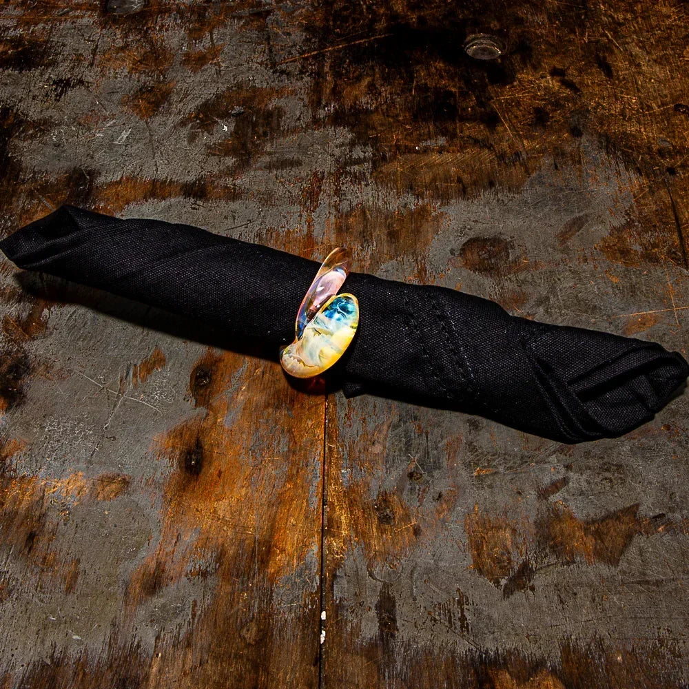 Handmade glass napkin ring with multicolor swirl on black cloth napkin, rustic wood table.