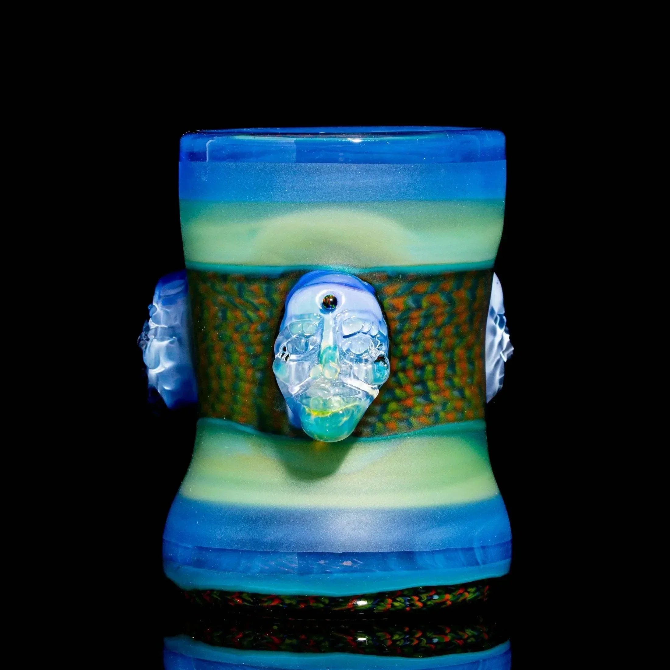Blue and green heady glass drinking vessel with sculpted face accents on black background.