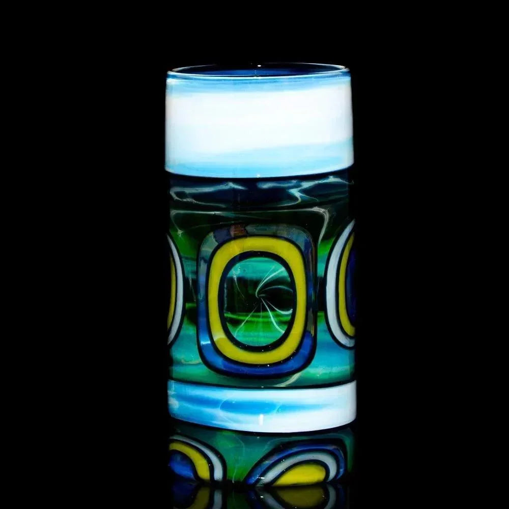 Hand-blown glass pint with blue, white, yellow, and green geometric patterns on black background.
