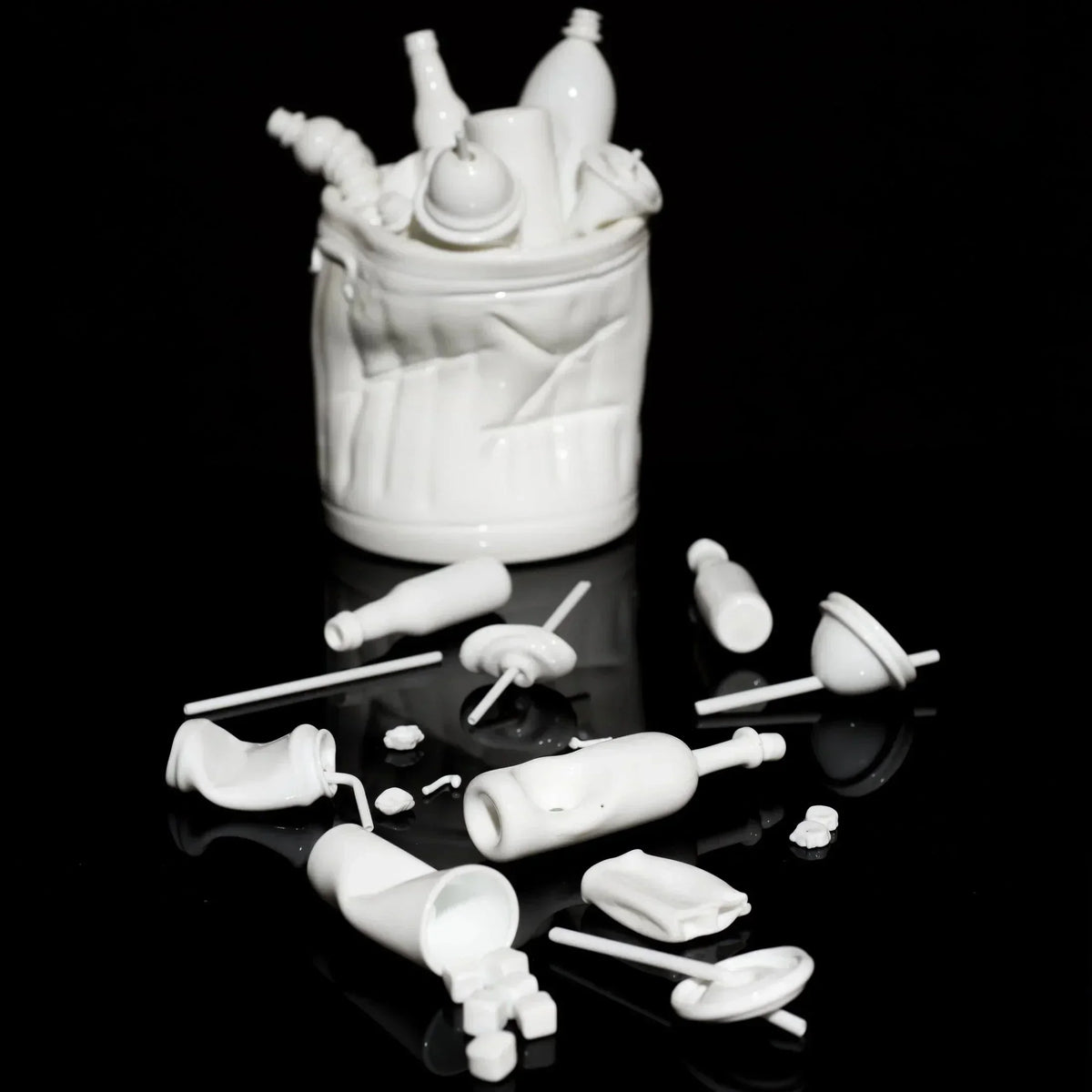 White ceramic art sculpture of a trash bin with bottles and scattered objects on a black surface