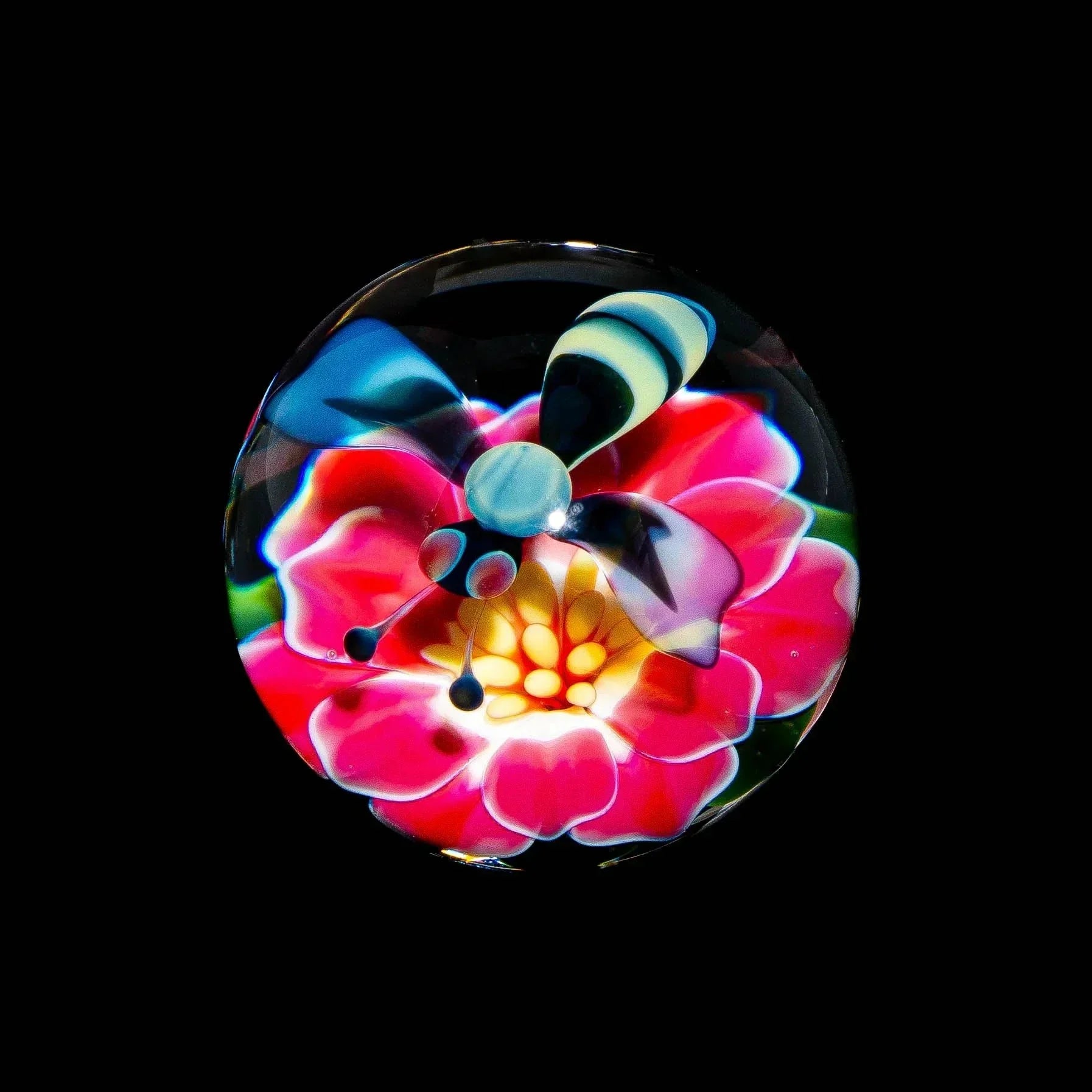 Handcrafted glass marble with a bee and vibrant pink flower design on a black background