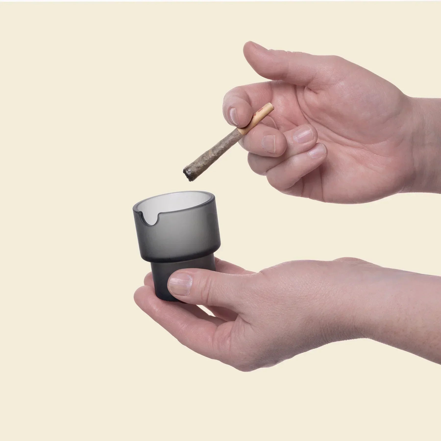 Hand holding a charcoal glass ashtray with a joint above it on a plain background