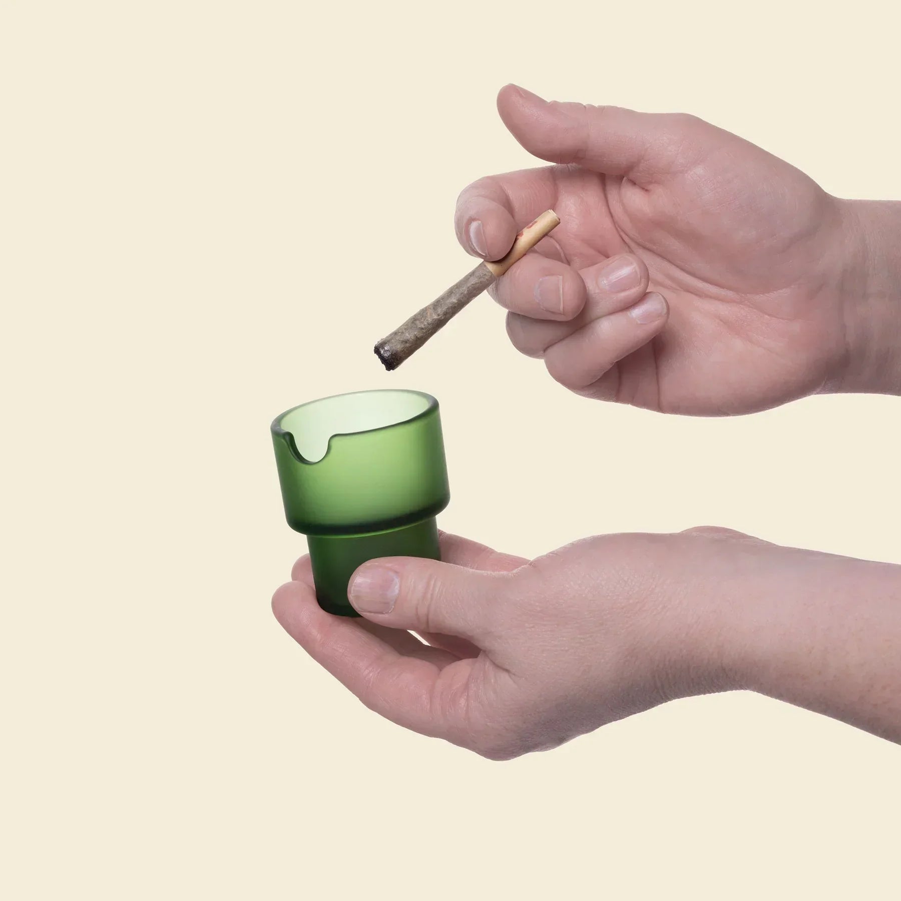 Hand holding green glass ashtray with joint above on light background