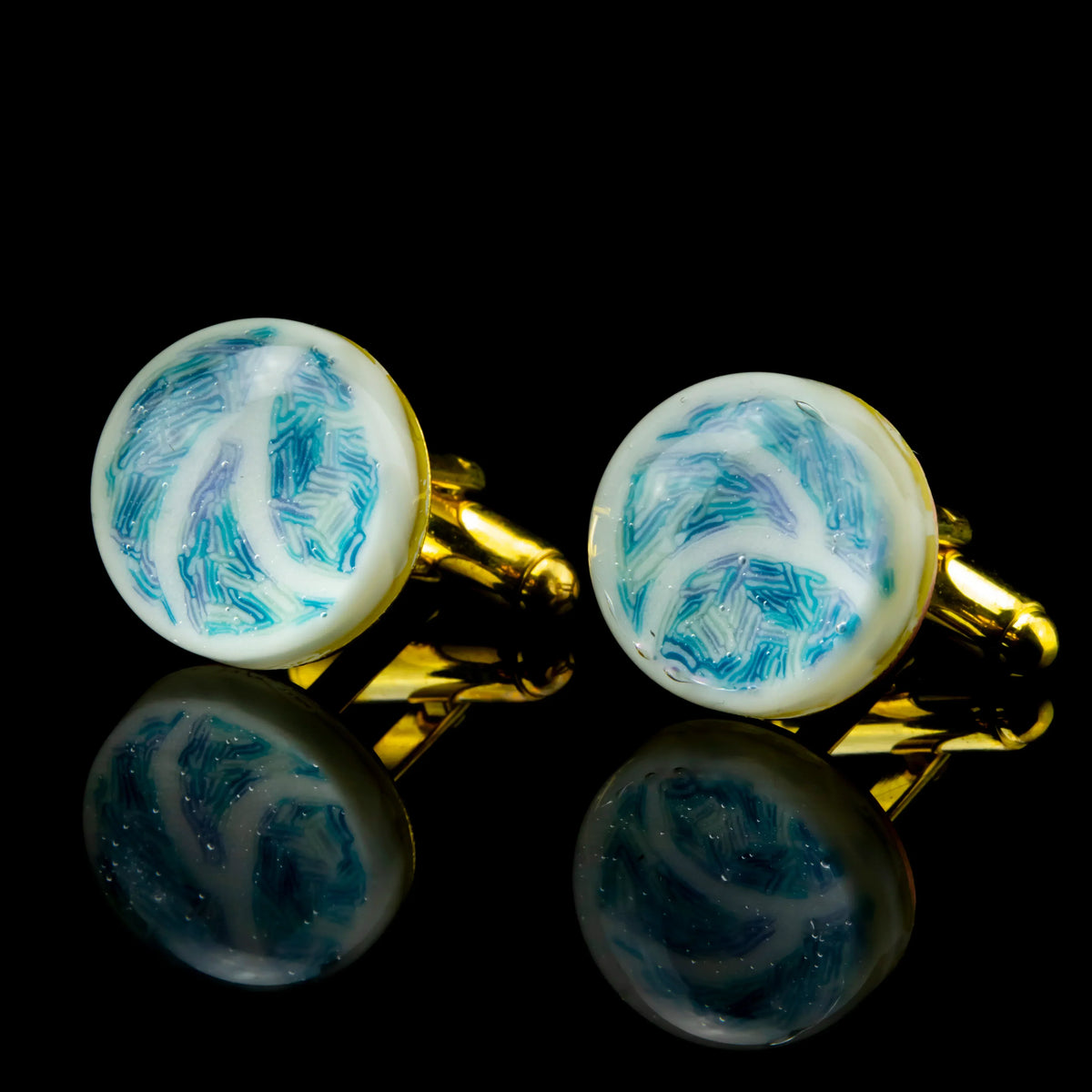 Round gold cufflinks with blue and white abstract pattern on a black reflective surface