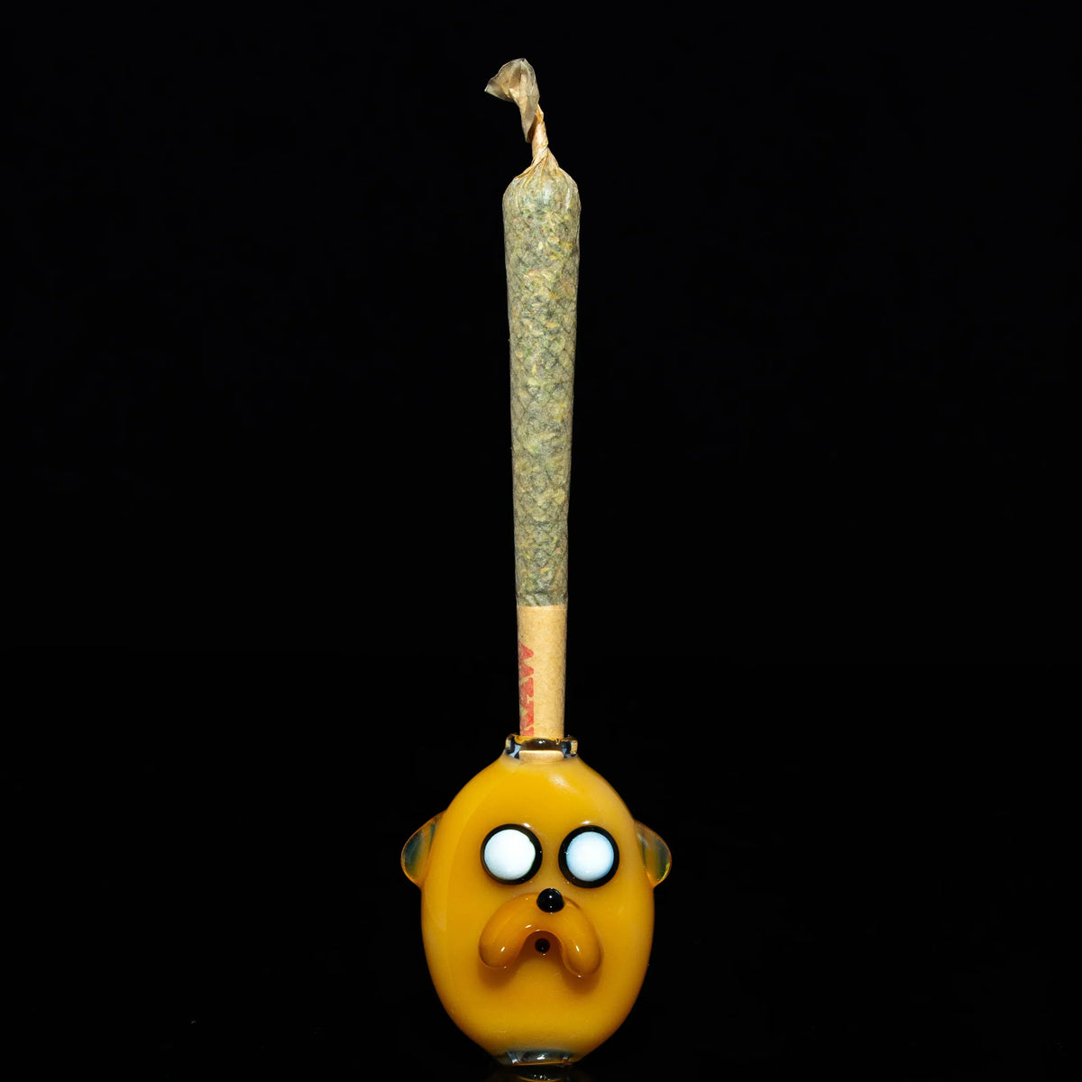 Hand-blown glass joint holder with cartoon face, holding a pre-rolled cone, on black background.