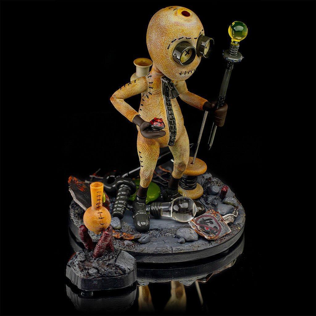 Heady glass art figure with stitched details, goggles, and staff on dark display base.