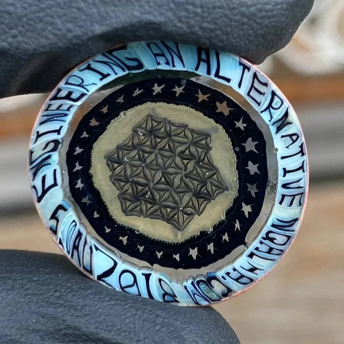 Glass marble with geometric pattern, star accents, and blue text border held by gloved hand