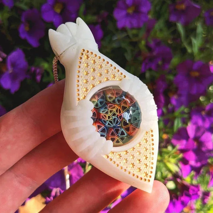 Hand holding intricate white glass pendant with colorful geometric center and gold accents, outdoors with purple flowers.