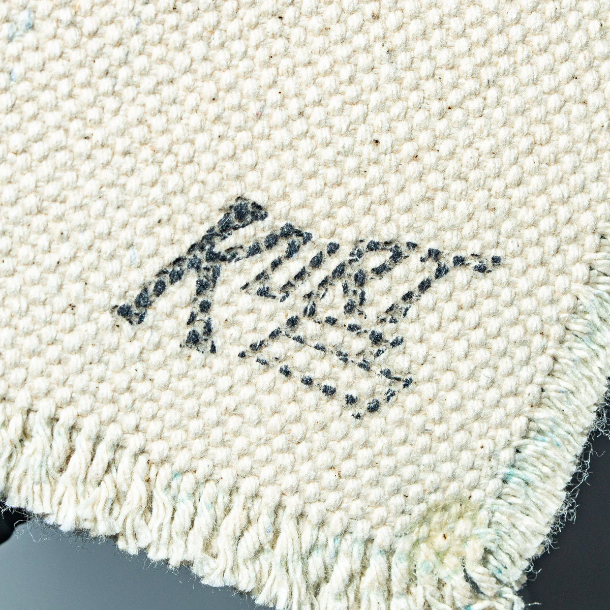 Close-up of handwoven cream glass mat with black artist signature and fringe edge