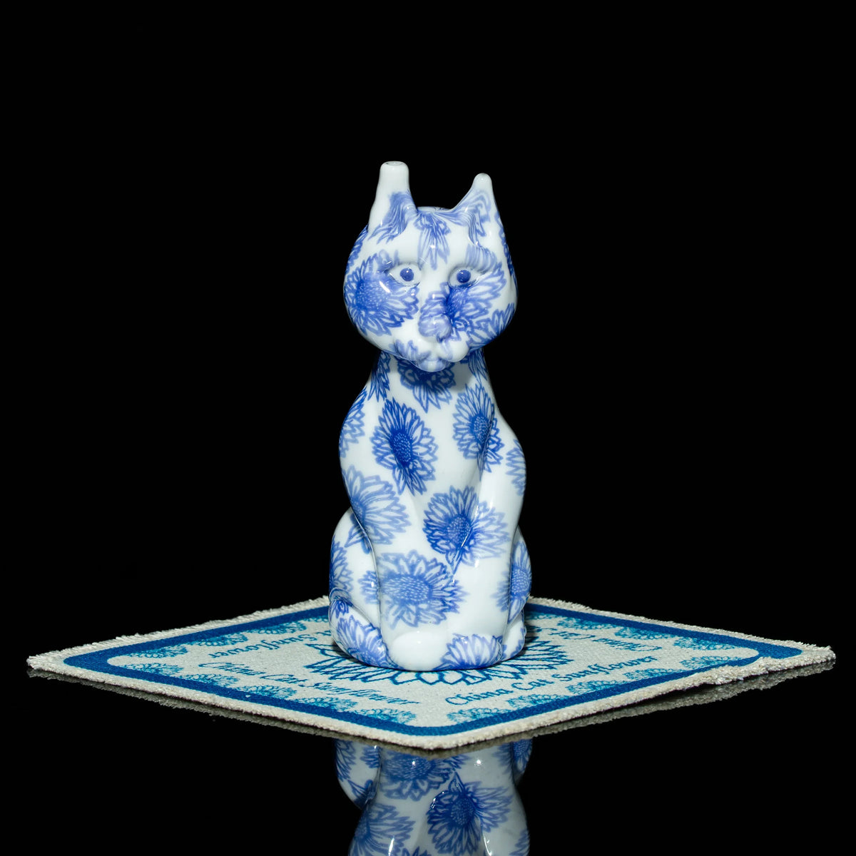 Blue and white floral glass cat figurine on a patterned coaster, black background