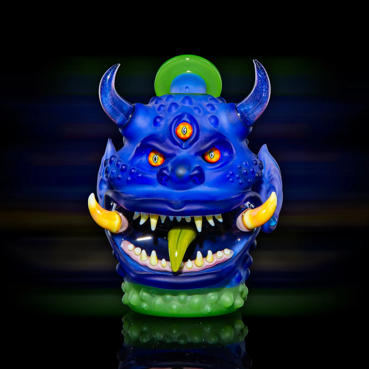 Colorful blue glass monster art piece with three eyes, horns, fangs, and green tongue