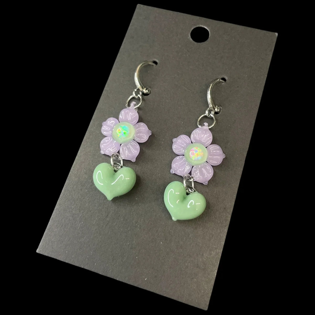 Handcrafted glass earrings with purple flower and green heart charms on black card