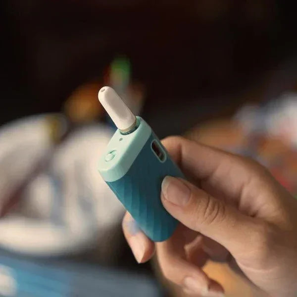 Hand holding a compact teal portable vaporizer with white mouthpiece indoors