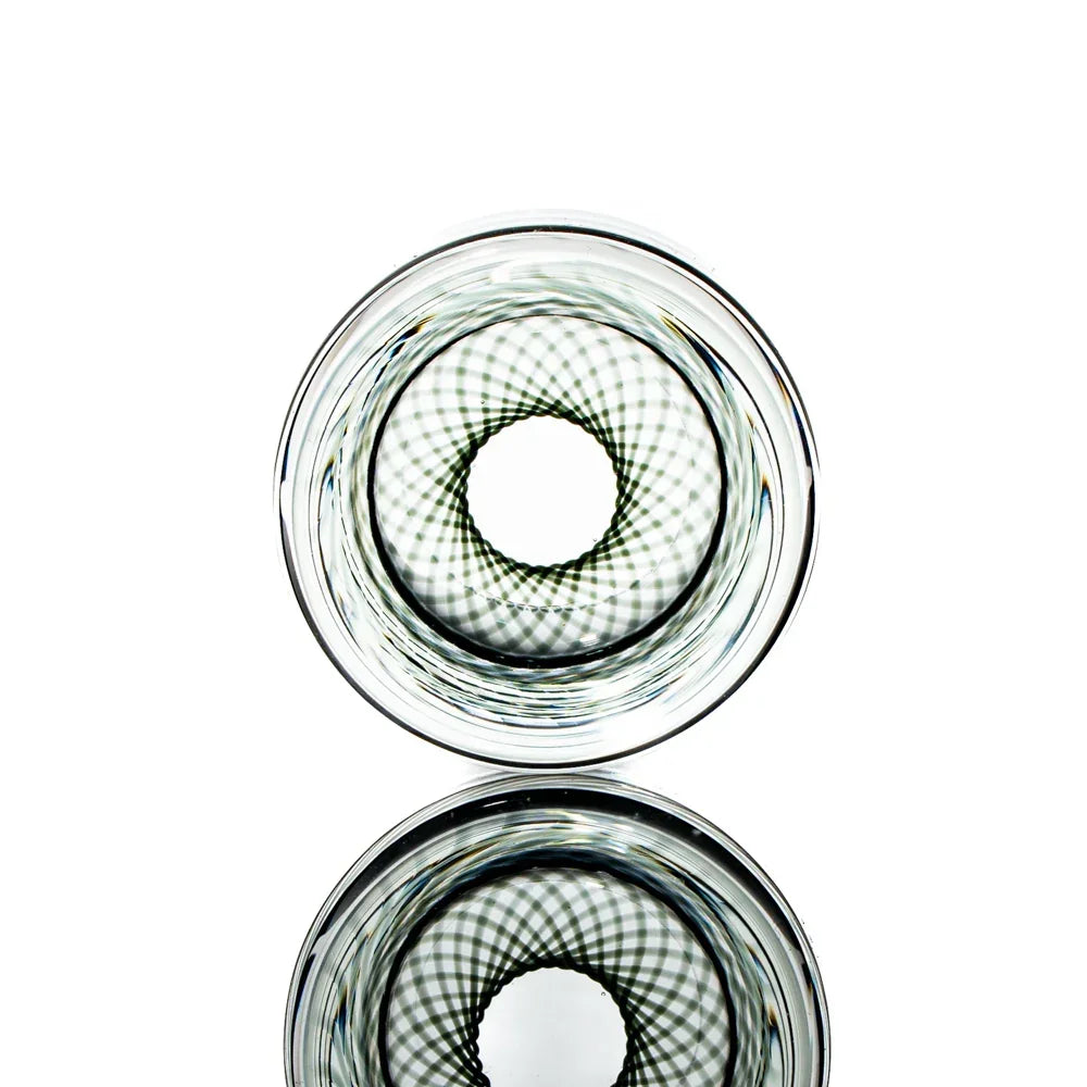 Clear glass shot glass with intricate black spiral pattern, reflected on white surface