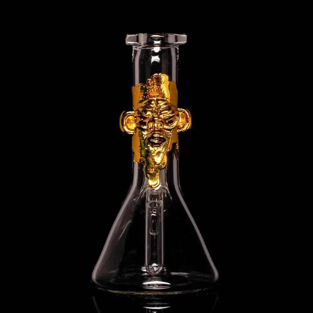 Clear glass beaker with gold face sculpture accent, displayed on black background