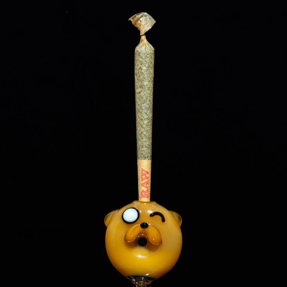 Cartoon dog glass joint holder with winking face, holding a rolled RAW cone against black background