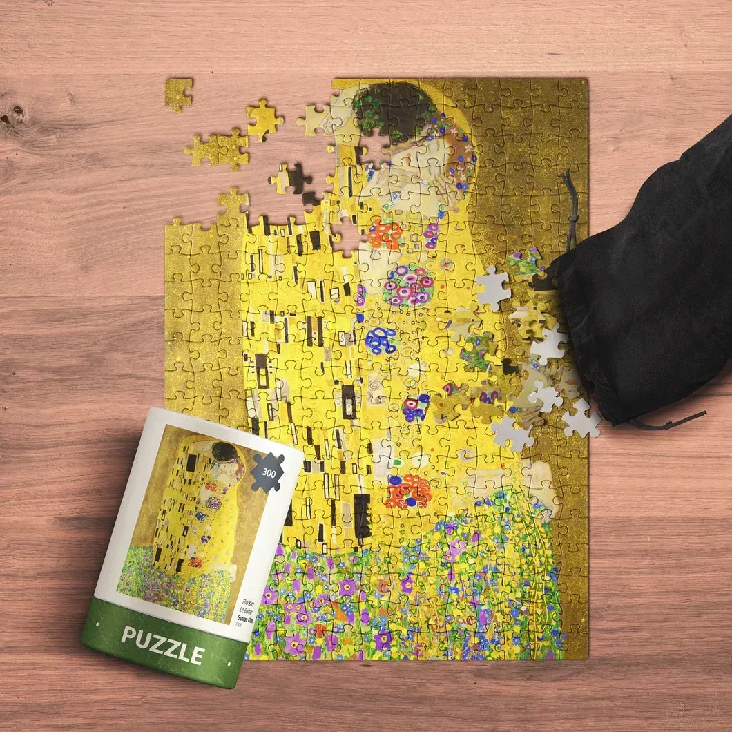 Partially completed art puzzle with yellow and floral pattern, puzzle box and black pouch on wood table