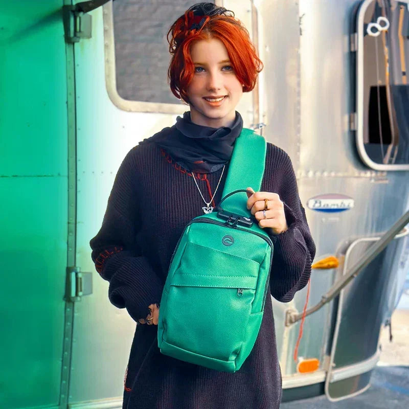 Person with red hair wearing black sweater and green sling backpack in front of trailer