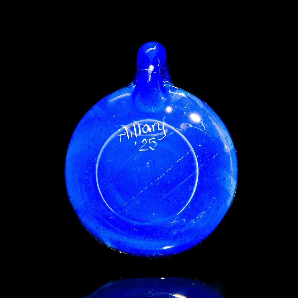 Blue glass pendant with a small loop, artist signature and year inscription, on black background.