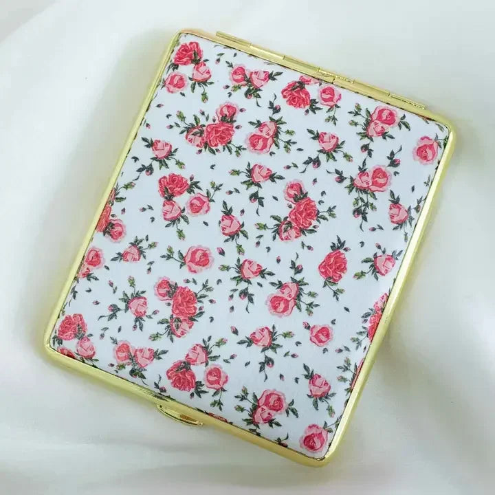 Floral metal case with pink rose pattern and gold trim, displayed on a white fabric background.