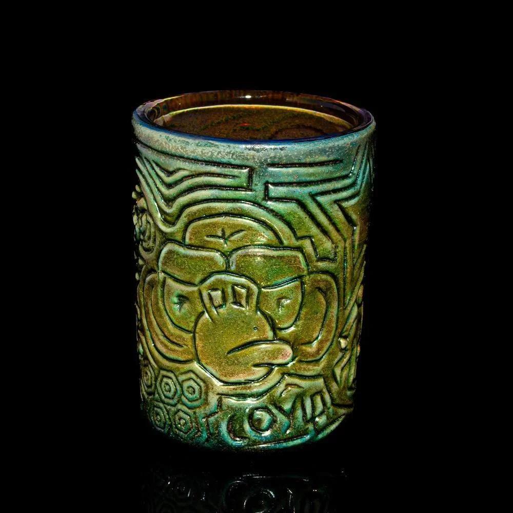Green ceramic shot glass with intricate embossed monkey face design, displayed on black background.