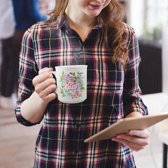 Woman holding floral ceramic mug with 'Stay High' design and reading tablet indoors