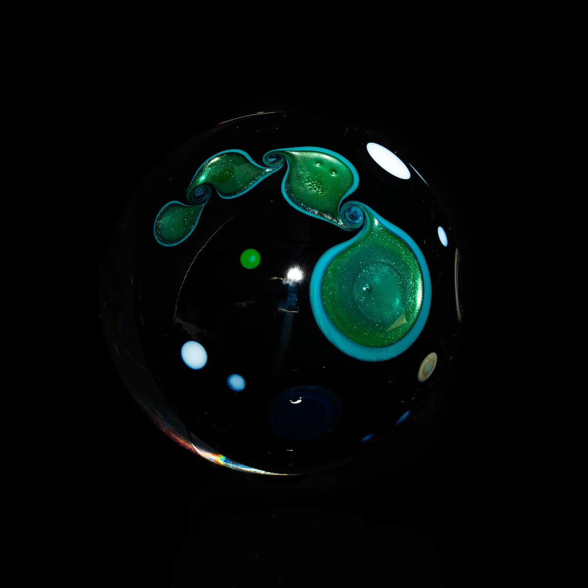 Hand-blown glass marble with green and blue swirling patterns and dots on black background
