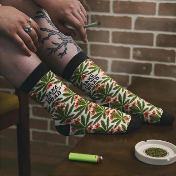 Person wearing colorful socks with cannabis leaf pattern and 'I take the high road' text, sitting by a table with a lighter and ashtray.