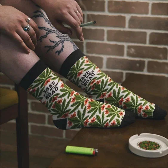 Person wearing colorful socks with cannabis leaf pattern and 'I take the high road' text, sitting by a table with a lighter and ashtray.