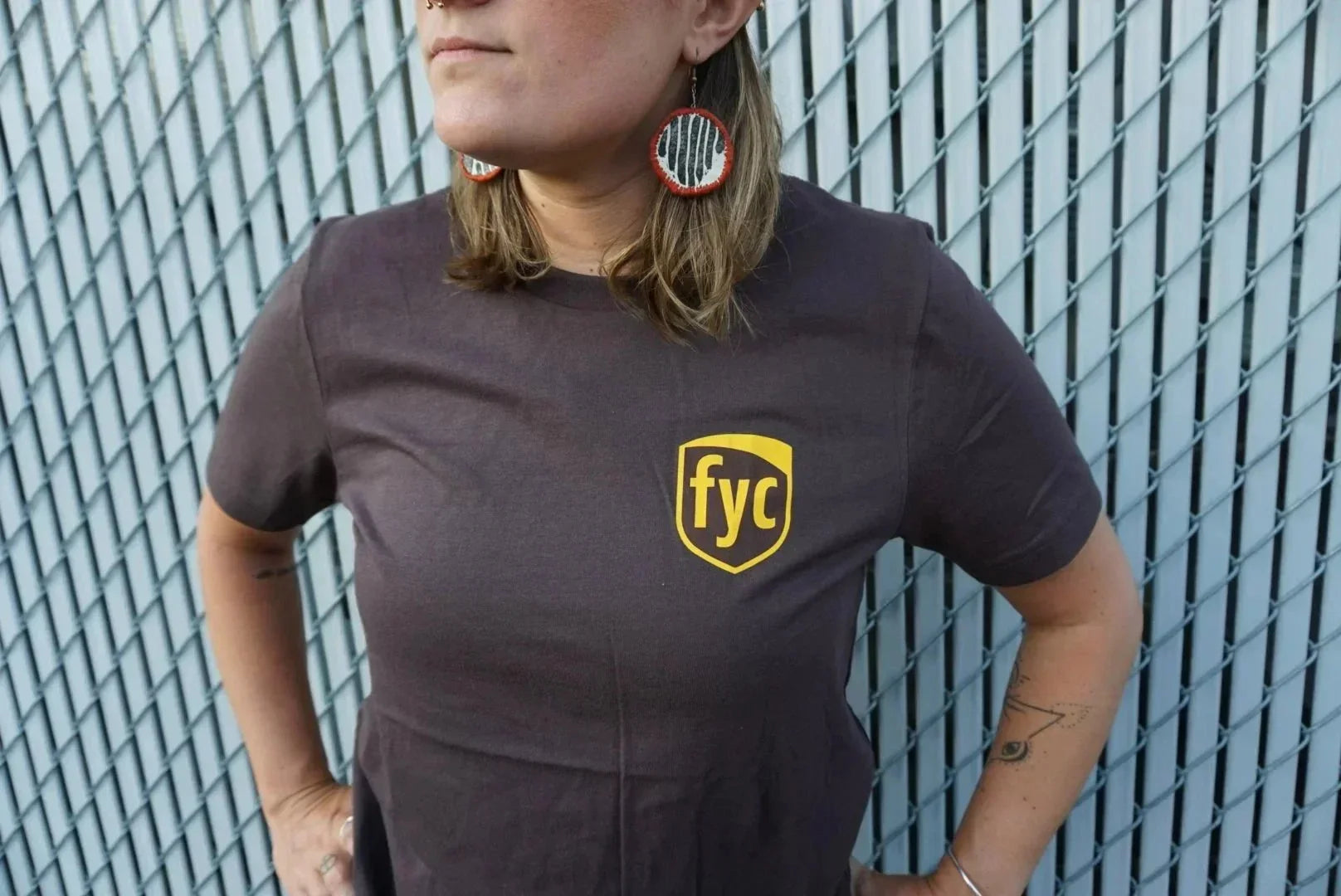 Person wearing dark t-shirt with yellow fyc logo, standing by chain link fence