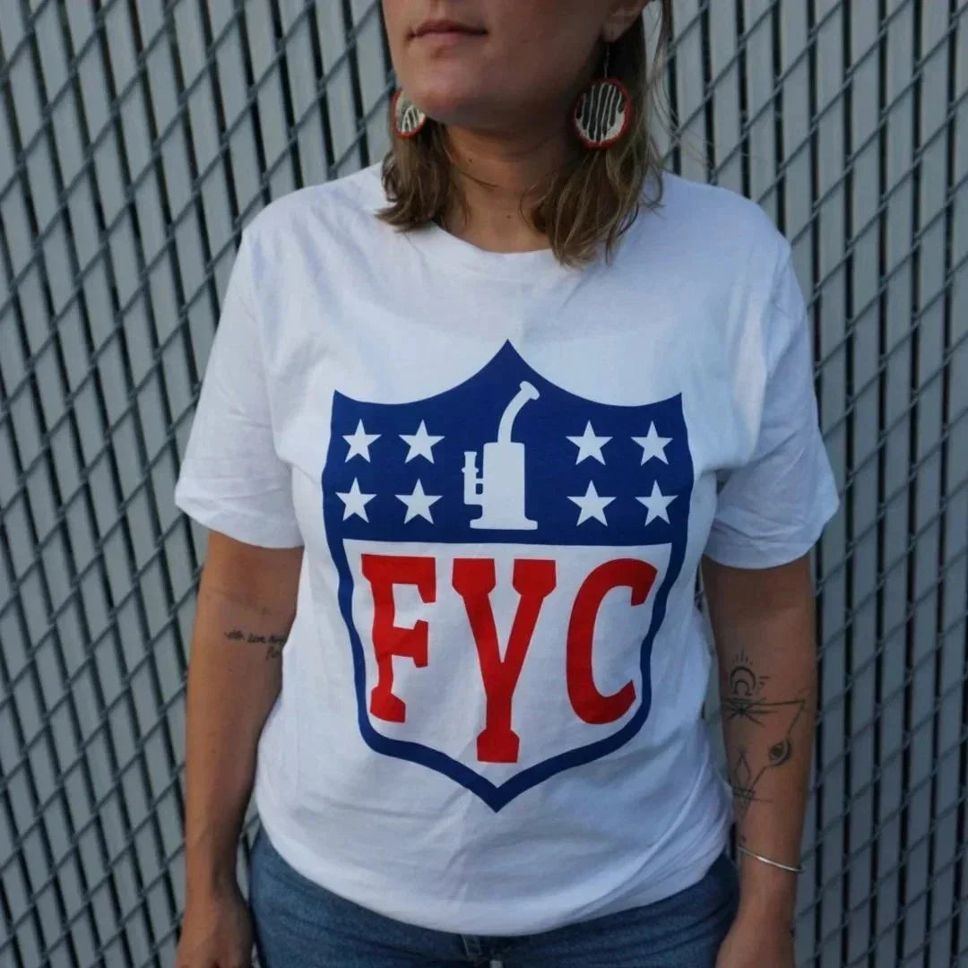 Woman wearing a white FYC t-shirt with dab rig graphic and stars, standing by metal fence.