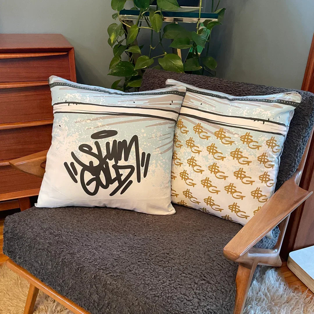 Two decorative throw pillows with graffiti and dollar sign designs on a mid-century chair